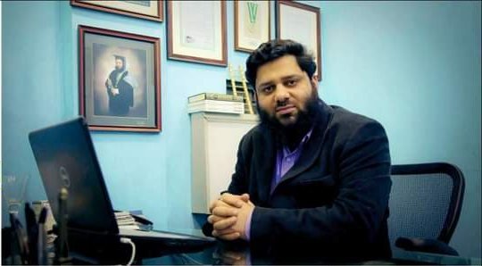 Dr. Ahmed Ejaz at his desk with framed portraits behind him